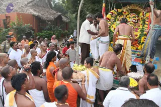 A temple procession on a village road beside a thatched-roof building. On a decorated wooden cart a deity murti is heavily garlanded with yellow and white flowers. Bare-chested priests in white dhotis with gold-bordered cloth work around the cart, one lifting offerings and another steadying the cart. Devotees in white and saffron-orange clothing surround it, some holding trays of bananas, coconuts, flowers and sweets. A uniformed police officer stands at left and a motorcycle is parked behind the crowd.