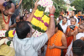A photograph of a temple procession outdoors. In the foreground a man in a grey shirt lifts a large garland of yellow and purple flowers with green leaves overhead. Behind him a man in an orange sleeveless robe with a gold bead necklace stands with hands folded, and beside him a man in orange with a camera rig and white dhoti holds a camera. A bare-chested priest with a red rudraksha mala and white sandal marks on his forehead stands at left. Devotees in white shirts and dhotis crowd around, and a child in an orange top stands at upper right. A temple gopuram with colourful sculpted figures appears in the background among trees.