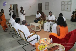 A photograph inside a bare, whitewashed hall with a concrete floor. A long-haired figure in a saffron-orange robe sits on a red velvet chair at the right, facing a semicircle of men seated on cane-backed chairs. Most men wear white dhotis and white shirts; one central man wears a white dhoti with a bare upper body and vibhuti on his forehead. A standing man in a saffron robe with a garland stands at left, and another in white holds a cloth at the back. A cameraman is visible in the upper left. On the low table in front are a plate of fruit, rudraksha malas, printed cards and a water bottle. A deity murti stands at the far right edge.