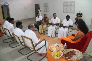 A seated group meeting in a plain whitewashed hall with a concrete floor. In the red-cushioned chair at right sits a person in orange robes with a white forehead mark and long dark hair, gesturing toward the group. Opposite and along the sides, about seven men sit on green-framed cane chairs, most in white shirts and white dhotis; one in the centre wears a saffron-yellow dhoti and bare upper body with a sacred thread and a white tilak, and another wears a white shirt with a tilak. A brass tray with rudraksha malas, sweets and offerings sits on an orange cloth, with a bottle of water, a basket of fruit, and pamphlets. A small dark deity murti stands on a shelf near the right wall.