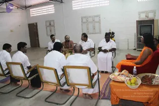A photograph of a seated group in a plain white-walled hall. A bare-chested man with a red-and-white shawl and rudraksha mala sits centrally in a cane-backed chair, facing the group. Several men in white shirts and white dhotis sit in a circle on an orange cloth. On the right, a person in an orange robe with long dark hair sits on a red chair, gesturing. A small table draped in orange holds a brass plate with fruit, sweets and a water bottle. A television and a small deity figure stand against the wall.