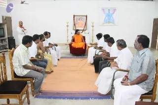 Indoor photo of a seated meeting in a plain white-walled room with a red-and-blue patterned rug. A man in saffron robes with a rudraksha mala and a long dark hairstyle sits on a red cushioned throne-like chair at the centre, hands resting on his lap. Around him, a semicircle of men sits on wooden chairs: several wear white dhotis and shirts, one wears a striped shirt and grey trousers, and one wears a light blue shirt, watch and gold bracelet. A shaven-headed man in white with an orange shawl stands at the left. Behind the throne hangs a framed portrait of a saffron-clad figure garlanded with red flowers, flanked by brass lamps. A large painting of a blue-skinned meditating figure hangs on the wall at upper right. A television stands at left.