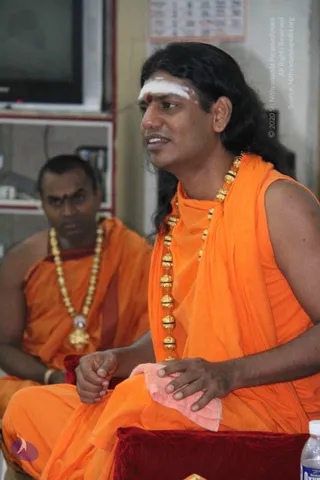 A photograph of a long-haired man in saffron-orange robes seated on a red velvet cushion, speaking while his hands rest on a pink cloth over his knee. He wears a white vertical forehead marking with a red dot and a long gold bead necklace. Behind him sits a second man in orange robes with a gold bead necklace and a gold pendant, also seated and looking on. A water bottle is visible at the lower right, and a wall calendar and shelving are in the background.