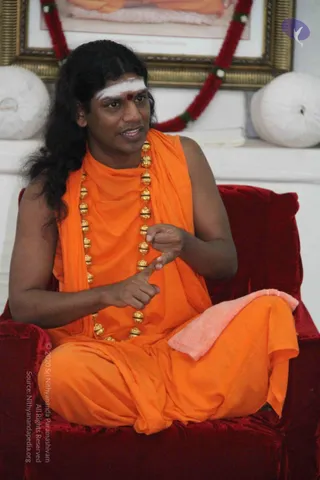 A photograph of a long-haired man seated cross-legged on a red velvet chair, speaking while gesturing with his hands. He wears a sleeveless saffron-orange garment draped over one shoulder and a long orange cloth, with a heavy necklace of gold beads with black bands and a white tilak with a red bindi on his forehead. A pink towel rests on his knee. Behind him are white bolster cushions, a gilded framed picture garlanded with red flowers, and a white wall. The man appears to be a probable Swamiji on the basis of his long swept-back hair, saffron clothing and gold bead necklace, and he is in_person.