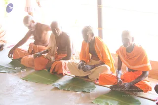 Four men sit cross-legged on a bare floor in bright daylight, with banana leaves and small plastic cups set in front of them. The man on the far left is bare-chested with a sacred thread and an orange cloth around his waist. The second man is bare-chested with a cream shawl draped over his shoulders and an orange lower cloth. The third wears an orange shawl over a bare chest and cream dhoti, and the man on the far right wears an orange shawl and orange dhoti, with his hands busy in front of him. All four have white ash marks on their foreheads and bodies. A pillar and a bright white wall stand behind them. Their pose and the leaf plates suggest a seated meal or offering, but the image alone does not show more.
