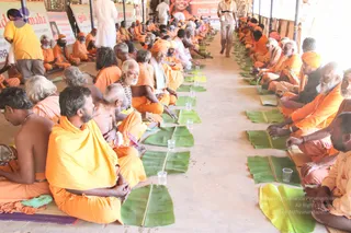 A photograph of a long covered hall where many saffron-clad sadhus and devotees sit cross-legged in rows on the floor for a meal. Each seated person has a fresh green banana leaf placed before them with small glass tumblers of water beside the leaves. Many men wear orange or saffron cloths, some with orange turbans and rudraksha malas, and several have long white beards. In the foreground a man in a yellow shirt is seated, while a man in white walks along the aisle between the rows. Banners with orange and red lettering hang on the wall behind the hall.