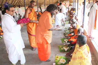 A large open hall with a thatched roof and bamboo-style ceiling where a line of seated people eat from banana-leaf plates arranged along the floor. A man in orange robes with long matted hair and a beard stands in the center serving food from a tray onto the leaves. Beside him a younger man in orange dhoti and shawl with a rudraksha-style necklace stands barefoot. On the left a man in white dhoti and shirt holds a tray of pink dragon fruit. Elderly seated devotees in orange and yellow cloth receive rice and side dishes, with white-clad attendants behind them and a small cup of water beside each plate.