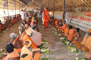 A long covered hall with a thatched roof and rows of seated sadhus and devotees in orange and saffron robes facing a central aisle, each seated before a plantain leaf with rice and food, cups of water nearby. A standing man in orange robes and a rudraksha mala holds a lit flame or aarti lamp at the centre of the aisle, while a man in white stands at the right. Banners behind show Tamil and Sanskrit script and a framed photo display. The scene resembles an annadan or prasadam meal offering with fire ritual.