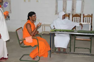 A photograph taken indoors in a plain room with white-washed walls, a concrete floor and wooden-backed chairs by a window. A man in an orange saffron robe sits on a cane-seated chair with a pink towel draped over its arm, gesturing with both hands as he speaks, with a white vibhuti mark on his forehead and a dark rudraksha-style bead necklace; his long dark hair is swept back. Across a metal-framed table sits a man in plain white dhoti and shawl with a white head covering, gesturing toward the table where two green banana leaves are laid out. A third man in white dhoti stands at the left edge holding a camera.