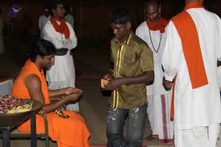 Photograph of an evening gathering on a plain floor at night. A seated man with swept-back dark hair wears an orange saffron cloth draped over his shoulders and bare arm, with a gold bangle on his wrist, and is handing a small item toward a tray of sweets and bananas on a table in front of him. A young man in a striped olive-and-yellow shirt and jeans stands before him holding an orange packet. Behind them, several men in white dhotis and orange shoulder cloths stand watching, one wearing a rudraksha mala, and a shaved-headed man in white stands on the right. The seated figure matches a Swamiji (probable) candidate by saffron clothing, swept-back hair and ceremonial gold adornment, seated and handing out offerings.