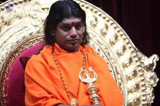 A man with long swept-back dark hair sits on an ornate gold throne with a pink cushion, wearing an orange saffron shawl over his shoulders and a rudraksha mala with a gold ceremonial pendant. He holds a gold trident-shaped staff upright in front of his body, with a red-brown tilak on his forehead. The background is a dark maroon curtain. Swamiji (probable) is seated centrally, identified by the saffron clothing, long hair, rudraksha mala and gold trident staff.
