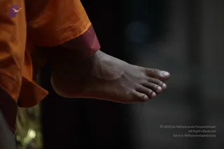 Close-up photograph of a bare foot and ankle of a person wearing orange and maroon robes, the foot raised slightly off the ground against a dark, blurred background. Warm orange fabric falls over the leg, and a faint golden object is visible at the lower left edge. Only the foot and lower garment are visible, so no face or other person can be seen.