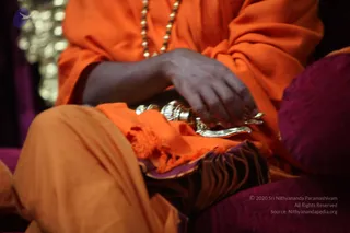 Close photo of a seated figure in saffron-orange robes and a matching orange shawl, with a strand of rudraksha-style beads around the neck. Their hands rest on a silver ritual object, possibly a bowl or vessel, on a draped orange cloth over a purple cushion. Only the torso, hands and lap are visible; the face is out of frame. Purple and gold drapery forms the background, with a blurred silver-toned object at upper left.