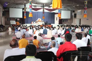 A wide photograph of a large indoor hall during a seated satsang. Dozens of devotees sit on red-and-white striped mats and rows of black chairs, many facing away from the camera toward a raised blue-curtained stage. On the stage, several people in white and saffron sit near a central seated figure in orange robes before a white draped backdrop, with a small ritual setup in front. Two projection screens flank the stage, and tall vertical banners with woven blue, red and yellow patterns and gold tassels hang from the white ceiling drapery. Men in white kurtas, orange and red shirts are visible in the foreground, and a single person in an orange robe stands to the far left near a whiteboard.