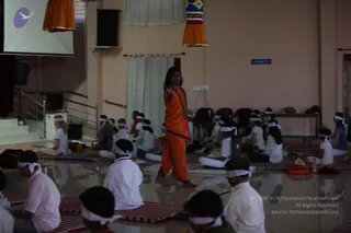 A photograph of a meditation or pranayama session in a hall with pale pink walls, a projector screen at left, and orange fabric hangings. A standing man in an orange saffron robe with a rudraksha mala draped over his shoulders walks between rows of seated participants. The seated participants wear white headbands and white or pink shirts, sitting on red-and-white striped mats. The standing man has long dark hair and a dark forehead mark, and appears to be teaching or guiding the session. Chairs line the back wall.