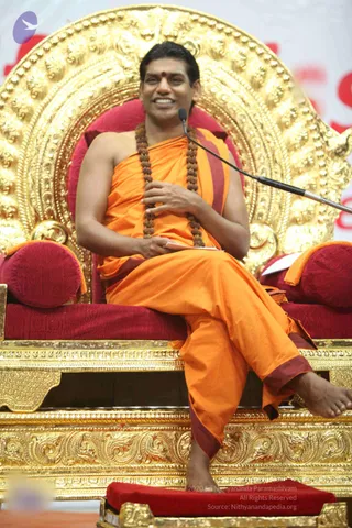 A seated man in an orange saffron robe with maroon borders sits on an ornate gold throne with a red velvet cushion and backrest. He wears a long rudraksha mala around his neck, a red bindi on his forehead, and holds a small white sheet of paper in his lap while a microphone stand curves in front of him. He smiles and speaks, his bare feet resting on a red footstool before the golden arched backdrop of a discourse stage. Swamiji (probable): long dark hair, saffron robe, rudraksha mala and a throne seating during a discourse.