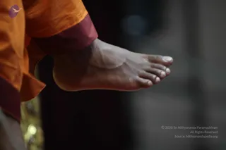 Close-up photograph of a bare foot and ankle of a person wearing orange and maroon robes, with the toes pointing right and the leg extended in a blurred dark background. The orange fabric drapes at the left edge, and a faint gold object is visible at the lower left. The foot is the sole visible body part.
