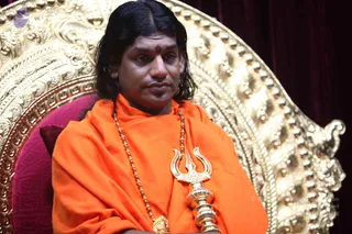 A man seated on an ornate gilded throne with curved scrollwork and studs, in front of a deep maroon curtain. He wears an orange-saffron shawl over his shoulders and a rudraksha mala with a gold pendant, and holds a gold trident-shaped staff across his chest with both arms folded. He has long dark swept-back hair and a red tilak on his forehead. Swamiji (probable) is a seated figure with long swept-back hair, saffron robes, a rudraksha mala and a ceremonial trident, seated on a throne.