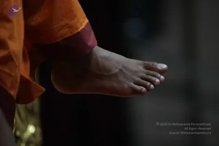 Close-up photograph of a bare foot lifted off the ground, with bare toes and a visible ankle. The leg is wrapped in orange saffron cloth with a deep maroon band at the ankle. The background is a dark, softly blurred interior with a brass or gold object at the lower left edge. The foot is the only visible body part, and no face is shown. The orange cloth and maroon trim are the strongest colours in the frame.