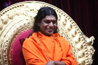 A seated man in an ornate gold throne with a pink cushion and deep red velvet drapery behind him. He wears a saffron-orange shawl wrapped over his body, a rudraksha mala of beads around his neck, and a red bindi on his forehead. His long dark hair is swept back and his arms are folded in his lap as he smiles. Swamiji (probable) appears to be the seated figure, based on the orange clothing, rudraksha mala and long hair.