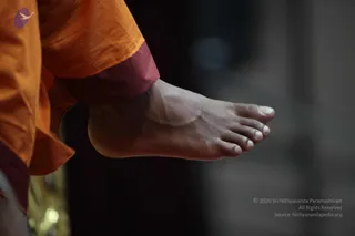 Close-up photograph of a bare foot and ankle of a person wearing saffron-orange robes with a maroon hem band at the wrist-length sleeve edge. The foot is lifted slightly off the ground with toes relaxed, against a dark blurred background with a soft grey light. Only the lower limb is visible; no face, head or other body parts appear in the frame.