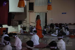 A photograph of a yoga or pranayama session in a bright hall with a projection screen at left and red curtains. A standing man with long, swept-back dark hair wears an orange saffron wrap, bare-chested, with a rudraksha mala across his chest, and gestures with one outstretched arm toward the seated group. Many participants sit on red-and-white striped mats wearing white clothes and white headbands; some sit on folded blankets and bags, and a woman in a light top sits near the front left. Orange-and-gold banners hang from the ceiling and a line of chairs runs along the right wall. Swamiji (probable): the standing figure has long hair, a saffron wrap and a rudraksha mala.