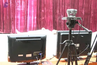 A photograph of a stage set-up in front of a draped magenta velvet curtain. In the foreground two flat-screen televisions are seen from behind, their back panels and cables visible, with a grey pan-tilt camera mounted on a black tripod between them. Behind the displays a white cloth covers the lower stage area, and a small printed item lies on the wooden surface at the bottom. No people are visible in the frame.
