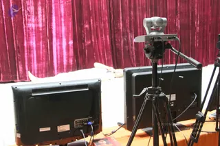 A photograph of studio-style equipment in front of a draped magenta velvet curtain. In the foreground, two black flat-screen monitors seen from behind face away from the viewer, with cables and connectors visible on the rear of the left screen. A silver pan-tilt camera sits on a black tripod at centre, beside a second tripod on the right, and an orange tablecloth covers the table. Behind the equipment, a person in white lies stretched out on a white surface at the far left, partly hidden by the screens. A small printed booklet or photo lies on the table at the lower left.