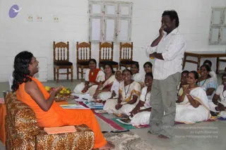 A photograph of a meeting in a plain whitewashed hall with wooden-framed windows and wooden chairs along the back wall. A figure with long swept-back dark hair, wearing a saffron-orange robe with a bare shoulder, sits on a leopard-print cushioned chair at left, gesturing with raised hands and a plate of fruit and flowers beside him. Around fifteen women seated on the floor on colourful striped mats wear white saris with gold borders, several with lanyard badges, and one wears an orange shawl. A standing man in a white striped shirt and grey trousers, hand raised to his face, speaks to the group. A small purple logo appears in the upper left corner.