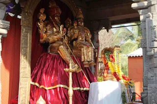 Golden murti statues of a deity couple seated together on a raised crimson satin platform under an ornate gold-carved arch within a stone temple pavilion. The male figure holds a staff and a ceremonial object in the raised hand, wears a red turban and golden armour-like ornaments; the female figure wears gold jewellery and holds a flower. Pink flowers adorn the figures. In the right foreground a cobra-hooded snake sculpture is draped with yellow and red marigold garlands and a strand of beads, beside a white cloth-covered table. Stone pillars with carved detail frame the scene and palm trees are visible outside. No people are present.