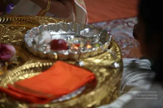 Close-up photograph of a ritual offering on a gold-toned tray draped with an orange cloth. A silver bowl holds small oil lamps, a red fruit, a white item and a pink flower. A bangled wrist in a white and orange sari is visible at upper left, and a partial profile of a person in white is at right. The setting appears to be a temple puja.