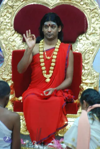 A seated man on an ornate gold-carved throne with a red velvet back and armrests raises his right hand in a mudra while his left hand rests in his lap. He wears a red draped garment with blue-edged borders, a heavy gold bead necklace and a gold chain, and has long dark hair with a red bindi and white markings on his forehead. Two people in white clothing sit in the foreground at the bottom corners, facing him, with their heads partly cropped, and pink flowers lie near the base of the throne. Swamiji (probable): the appearance cues of long swept-back hair, ornate gold necklaces and a saffron-toned draped garment on a throne fit the page's candidate, presented here in a satsang-style setting.