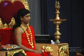 A seated man in a red wrap sits on an ornate red and gold throne at a satsang or discourse. He wears a heavy gold bead necklace and has long swept-back dark hair; his hands rest folded in his lap. Beside him stands a tall brass oil lamp with a lit flame and a bird-shaped finial, and a microphone and water bottle sit on a wooden podium. A dark blue curtain backs the scene, and a tray of offerings is at left. Swamiji (probable): long swept-back hair, red ceremonial wrap and gold ornate necklace on the throne. Two partially visible people sit at the lower edge of the frame.