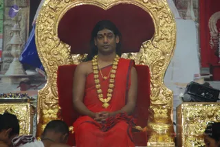 A man seated on an ornate gilded throne with a deep red velvet back and cushion, hands folded in his lap. He wears a red draped garment with dark blue trim and a heavy gold bead necklace, with a red bindi on his forehead and long dark hair. Swamiji (probable) based on the long swept-back hair, gold necklace and red-and-gold regalia on a throne. Foreground shows the heads of two seated devotees, and a gold-and-blue tray of flowers sits at left. Behind the throne are decorative red and gold hangings and a brass lamp. A small black audio device with microphone sits at right.