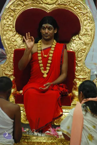 A man with long dark hair and a red bindi sits on an ornate gold-and-red throne, wearing a red robe with blue-edged borders and a heavy gold bead necklace with a gold pendant. He raises one hand in a mudra and rests the other in his lap. Two people in white garments are seen from behind in the foreground, and a few small incense sticks and pink flowers lie near the base of the throne. Swamiji (probable): the long hair, red robe and gold necklace match the page's description, and he is identified by role as the speaker seated on the throne.