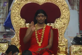A man seated on an ornate gilded throne with a deep red velvet back and seat raises his right hand in a blessing gesture. He wears a red saffron-style garment with blue border, a red bindi on his forehead, a long gold rudraksha-style mala and a heavy gold necklace. Long dark hair falls to his shoulders. Blurred heads of seated devotees appear in the lower foreground, and gilded ritual vessels and flower garlands sit beside the throne. A backdrop of gold and white ritual decor surrounds the scene. a person is seated in the central role, presiding over a satsang.