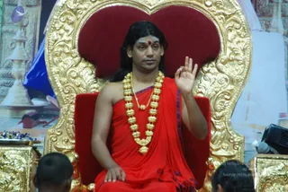 A man with long dark hair and a red bindi sits on an ornate gold and red throne, raising one hand in a teaching gesture. He wears a red shawl with blue edging and a thick gold necklace of beads with a pendant. Two partially visible heads of seated listeners appear at the bottom edge, and a brass oil lamp and a tray of blue flowers sit at the left. A backdrop with temple-style artwork fills the background.