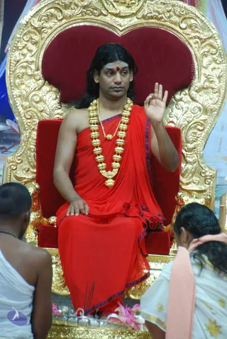 A seated man in a red draped garment with blue-edged border sits on an ornate gold-and-red throne. He wears a heavy gold bead necklace with a pendant, a red bindi on his forehead, and raises one hand in a blessing gesture while the other rests on his lap. In the foreground, two people in white clothing are seen from behind, one with a pink cloth draped over the shoulder. Incense sticks and pink flowers sit near the base of the throne.