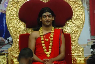 A man with long dark hair sits on an ornate gold-carved throne with a red upholstered back, wearing a red sash-like cloth draped over his bare shoulder, a red-brown bindi on his forehead, and a heavy gold multi-bead necklace with a smaller gold pendant. His hands are folded in his lap. In the foreground at lower left, the shaved head of another seated person is partly visible. Behind the throne, white and blue fabric hangings and a red curtain frame the stage.