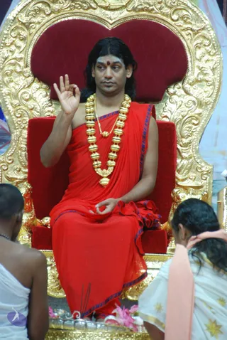 A man with long dark hair and a red bindi sits on an ornate gold-carved throne with red velvet cushions, wearing a red robe with blue trim and a heavy gold bead necklace. He raises his right hand in a mudra while his left hand rests in his lap. Two people in white garments with their backs to the camera sit in the foreground, and incense sticks and flowers rest on the platform below. A visible purple logo appears at lower left. The appearance-based label is probable Swamiji: the seated figure has the long hair, gold ceremonial necklaces and red robe associated with the page's named teacher, but the face is not enough for a firm identification.