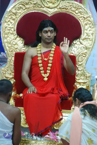 A man with long dark hair and a red bindi sits on an ornate gold-carved throne with red velvet upholstery, wearing a red draped garment with blue trim and a heavy gold bead necklace. He raises one hand in a teaching gesture while looking to the side. Two people in white clothing, seen from behind, sit below the throne in front of a small offering with pink flowers and incense sticks.