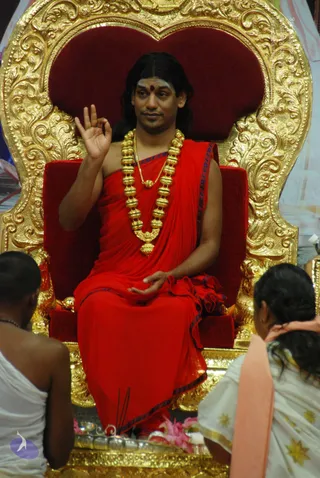 A man with long dark hair and a red bindi sits on an ornate gold-carved throne with red velvet upholstery, dressed in a flowing red robe with dark blue edging and a heavy gold bead necklace. He raises one hand in a mudra while resting the other in his lap, appearing to address the gathering. Two seated people in white garments face him from the lower foreground, one with an orange shawl and hair pulled back, the other partly turned away. Incense sticks rise near the base of the throne beside pink flower offerings. The setting is a satsang-style hall with draped fabric in the background.