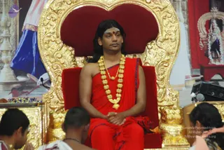 A seated man in red robes sits on an ornate gold-carved throne with a deep red cushioned back at a satsang setting. He wears a red shawl draped over one shoulder with blue-edged trim, a long gold necklace of ribbed beads and a red bindi on his forehead. His hands rest folded in his lap. Two people in the foreground, partly visible, sit with heads bowed, one in white with a rudraksha-style strand. Behind him a large painted backdrop shows a figure in blue and white and a red curtain with a white temple-style shrine on the right. Appearance-based label: a person, wearing red robes, a gold bead necklace and a seated throne posture.