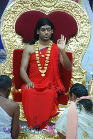 A seated man on an ornate gold-carved throne with a red cushioned back, wearing a red draped garment with blue-edged border and a heavy gold bead necklace with pendant. He has a red bindi on his forehead and long dark hair, and raises one hand in a teaching gesture. Two devotees in white stand or kneel in the foreground, one with a pink shawl, and incense sticks are visible near the base of the throne with pink flowers and a silver vessel.
