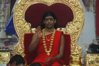 A photograph of a man seated on an ornate gold throne with a deep red cushioned seat and back, raising his right hand in a blessing gesture while his left hand rests in his lap. He wears a red saffron-style robe with a dark blue-and-gold border, a heavy gold multi-bead mala necklace, and a gold pendant, with a red bindi on his forehead. Partly visible at the bottom edge are the heads of two other people, and a microphone and camera equipment sit at the right. Behind the throne are gold-patterned hangings and a brass kalasha-style lamp stand at left. Swamiji (probable): the seated man has long swept-back hair and a heavy gold mala with a red robe, consistent with the page subject, though appearance is the only basis for this label.