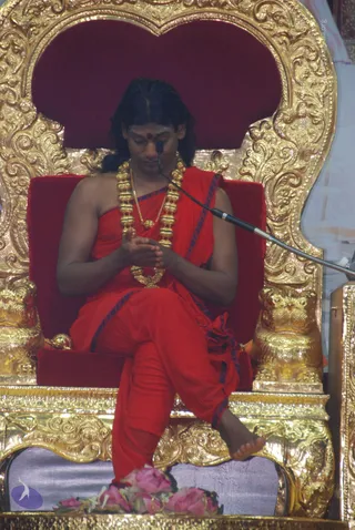A man with long dark hair sits on an ornate gold throne with a red velvet back and seat, eyes lowered and hands joined at his chest. He wears a red robe with blue trim draped over one shoulder, a red dhoti-style lower garment, a dark red bindi on his forehead, and a heavy gold necklace of beads with a pendant. A microphone on a stand is near his face. Pink roses lie at the base of the throne, and a small purple logo sits at lower left. The outline and bindi are consistent with a saffron-clad guru, but the figure is shown on a throne and the appearance cues are only partly distinctive, so identity is uncertain.