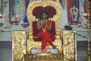 A seated man in a red saffron-toned robe sits cross-legged on an ornate gold throne with a red velvet seat and back. He holds his palms together in namaste at chest height with eyes closed, wearing a long gold-and-white beaded necklace and a red bindi. A microphone stand stands beside him and a black speaker sits near the throne arm. Pink flower petals lie on the throne base, and an ornate brass lamp stand with lit oil lamps appears at the right, with a painted temple backdrop behind. A figure in red stands in the background.