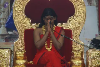 A man seated on an ornate gold-and-red throne with carved gilt scrollwork and lion-head armrests, hands pressed together in namaste at his chest with eyes closed. He wears a red robe with dark blue-striped edges, a red bindi, and a heavy multi-strand gold necklace with gold beads and a pendant. A microphone on a flexible stand is positioned before him, and a dark speaker or device sits at the right edge. The backdrop shows a large painted or printed banner and ceremonial objects. a person: long dark swept-back hair, ornate gold necklace and red robe, seated on a throne as the speaker in a satsang.
