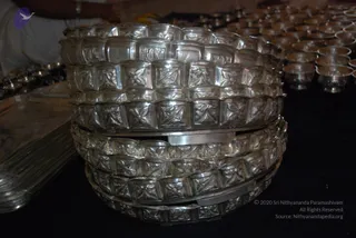 A stack of ornate silver-toned vessels with repeating embossed floral and geometric patterns sits on a black cloth in the foreground. Behind it, rows of smaller silver cups and bowls are arranged on dark fabric, and a silver tray lies at the left edge. No people are visible in the frame.
