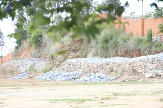 A photograph of a construction site beside a red brick boundary wall. A sloped embankment of exposed reddish-brown soil and scrub vegetation rises behind piles of grey cut stone and loose rubble laid along the foot of the slope. Bare sandy ground with sparse grass fills the foreground. Tree branches frame the top left. No people are visible.